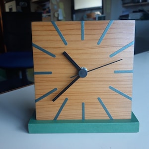 May include: A square wooden clock with a light brown face and teal lines indicating the hours. The clock has black hands and a green base. The clock measures approximately 15 cm wide and 15 cm tall.