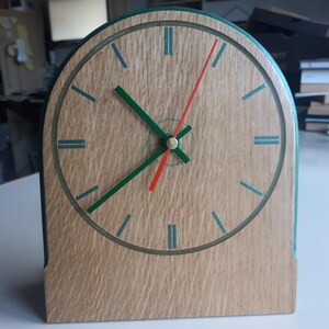 May include: A wooden desk clock with a light brown face and a green border. The clock features green and red hands, and blue-green hour markers. The clock is rectangular with an arched top.