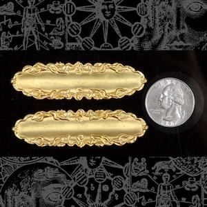 May include: Two gold-colored metal plaques with an ornate, floral design. The plaques are oval-shaped and have a smooth, polished surface. A silver quarter is included for scale.