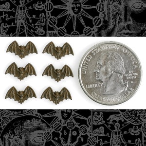 May include: Six small, bronze-colored bat charms, each with a slightly different wing shape. The charms are arranged in two rows of three, with a quarter dollar coin for scale.