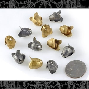 Puede incluir: Una colección de doce pequeños abalorios de metal en forma de flor, en tonos plateados y dorados. Los abalorios están dispuestos sobre una superficie blanca con una moneda de 20 céntimos de euro plateada para la escala.