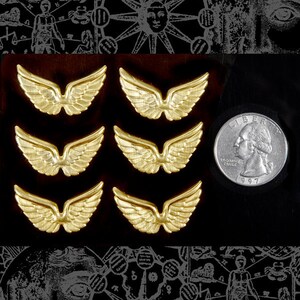 May include: Six gold-colored metal angel wings, each about the size of a quarter, are arranged in two rows of three. The wings are detailed and have a slightly textured surface.