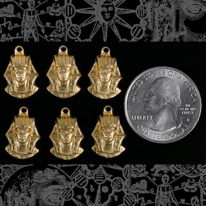 May include: Six gold-tone pendants featuring a profile of an Egyptian pharaoh with a headdress. The pendants are arranged in two rows of three. A US quarter dollar coin is included for scale.