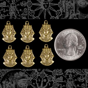 May include: Six gold-toned pendants featuring an Egyptian pharaoh bust design. The pendants are arranged in two rows of three. A US quarter is included for size comparison.