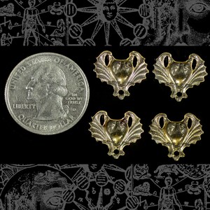 May include: Four gold-colored metal pendants with a bat-like face design, next to a U.S. quarter. The pendants have a detailed, textured appearance. The background is black with faint, stylized illustrations.