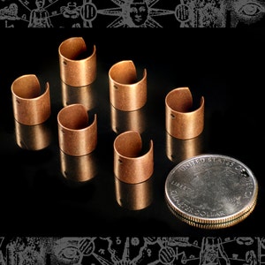 May include: Six copper ear cuffs, each with a small hole, arranged on a black surface. A US quarter dollar coin is placed below the cuffs for size comparison.