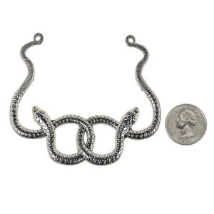 Op de afbeelding: Een zilverkleurige ketting met twee in elkaar verstrengelde slangen. De slangen zijn gedetailleerd met schubben en hun hoofden zijn naar buiten gericht. Een munt ligt ernaast als referentie voor de grootte.