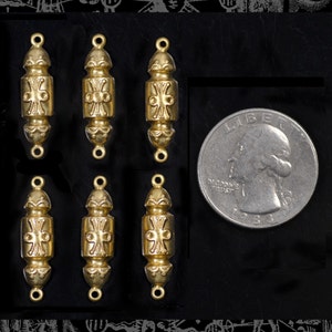 May include: Six gold-colored metal decorative beads with an ornate design, featuring a central cross-like pattern. Each bead has a loop at both ends. A U.S. quarter is included for size comparison.