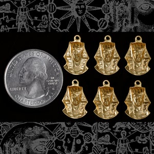 May include: Six gold-coloured pendants featuring an Egyptian pharaoh head design. The pendants are all the same size and shape, and they are arranged in two rows of three. The pendants are on a black background.