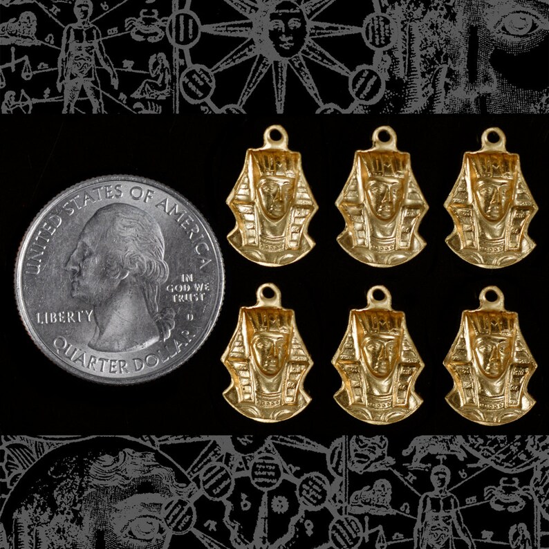 May include: Six gold-coloured pendants featuring an Egyptian pharaoh head design. The pendants are all the same size and shape, and they are arranged in two rows of three. The pendants are on a black background.
