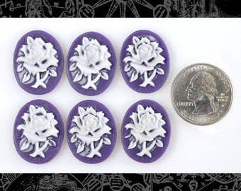 May include: Seven oval-shaped purple cameos with white rose designs. A U.S. quarter coin is next to the cameos for size comparison. The cameos are likely for jewelry making or crafting.