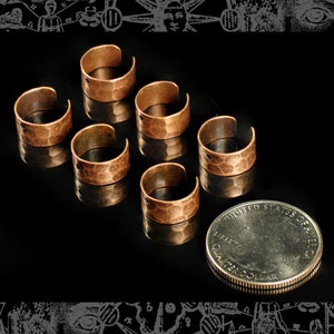 May include: Seven hammered copper ear cuffs, each with a small hole, arranged on a black surface next to a US quarter.