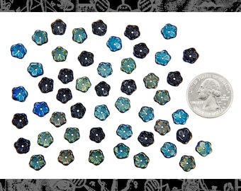 Puede incluir: Un conjunto de pequeñas cuentas en forma de flor en tonos azules, turquesas y negros, dispersas sobre una superficie blanca. Se incluye una moneda para referencia de tamaño. Las cuentas tienen un brillo metálico y un diseño de cinco pétalos.