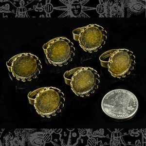 May include: Six gold-toned metal rings with a scalloped edge and a round, empty bezel. The rings are arranged on a black surface with a US quarter for scale.