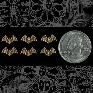 May include: A close-up of six small, gold-colored bat charms. The bats are arranged in two rows of three. The bats are all facing the same direction. The bats are on a black background. A silver quarter dollar coin is in the background.