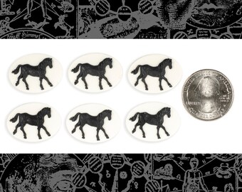 Könnte beinhalten: Sechs weiße, ovale Cabochons mit schwarzen Pferdesilhouetten. Die Cabochons sind in zwei Reihen zu je drei angeordnet. Eine US-Viertel-Dollar-Münze dient als Größenvergleich.