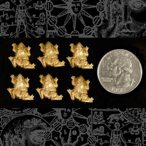 May include: Six gold-colored frog figurines on a black background. The frogs are facing forward with their legs outstretched. The frogs are about 1 inch in size.