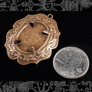 May include: A brass pendant with an ornate oval frame and a blank center. The pendant is about the size of a US quarter.