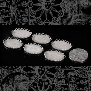 Puede incluir: Seis piezas ovaladas de metal plateadas con bordes festoneados. Las piezas est&aacute;n dispuestas en semic&iacute;rculo sobre una superficie negra. Una moneda de 25 centavos de d&oacute;lar estadounidense plateada se encuentra en la esquina inferior derecha de la imagen.