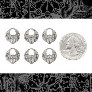 Puede incluir: Seis abalorios de escarabajo de metal plateado con alas detalladas y un lazo en la parte superior. Se incluye una moneda para la comparación de tamaños. Probablemente para la fabricación de joyas o manualidades.