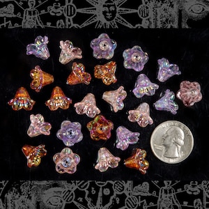 Può includere: Una collezione di perline a forma di fiore in vetro iridescente in tonalità di rosa, viola e arancio. Le perline sono disposte su una superficie nera e hanno diverse forme e dimensioni.