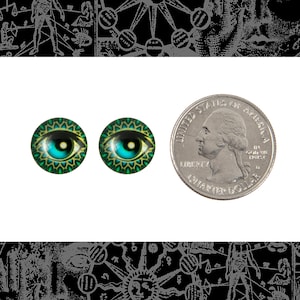 May include: Two round, teal and gold eye cabochons with a quarter for size comparison. The eyes have a detailed iris and pupil design. The cabochons are set against a white background with a black and white illustration.