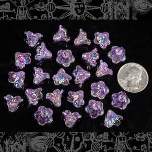 Può includere: Una collezione di perline di vetro a forma di fiore di colore viola iridescente. Le perline sono sparse su una superficie nera e hanno circa le dimensioni di una moneta da 25 centesimi di dollaro americano.