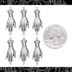 Puede incluir: Seis colgantes plateados con forma de mano, con diseños detallados y bucles para sujetar. Se incluye una moneda de 25 centavos estadounidense para comparar el tamaño. Los colgantes tienen una estética vintage o gótica.