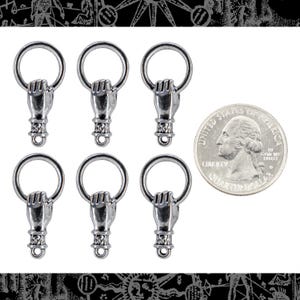 May include: Six silver-colored metal charms, each featuring a hand clenching a ring. A U.S. quarter coin is included for size comparison. The charms have a detailed, vintage appearance.