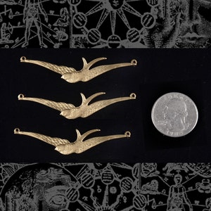 Puede incluir: Tres abalorios de metal de color dorado en forma de golondrina con bucles en cada extremo. Los abalorios están dispuestos en fila, con el abalorio superior ligeramente más alto que los demás. Se muestra una moneda de un cuarto de dólar estadounidense para comparar el tamaño.