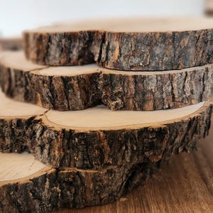 Puede incluir: Una pila de seis rodajas de madera natural con bordes de corteza rugosos y texturizados. Las rodajas de madera son de color marrón claro con un patrón de vetas visible, creando una estética rústica y orgánica. Las rodajas están apiladas unas sobre otras.