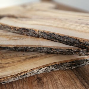 May include: Three wooden serving boards with natural, live edges are stacked on a wooden surface. The boards have a light brown colour with darker brown bark edges. The wood grain is visible, adding to the rustic aesthetic.