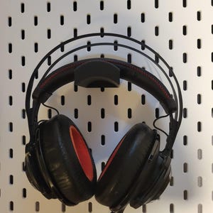 Könnte beinhalten: Schwarzer Over-Ear-Kopfhörer mit roter Innenpolsterung und schwarzem Kopfband mit roten Nähten. Der Kopfhörer hängt an einer weißen Lochwand mit schwarzen, ovalen Löchern. Ein schwarzes Kabel hängt herunter.
