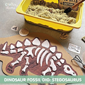 Könnte beinhalten: Ein gelber Kunststoffbehälter mit Sand, einer Schaufel mit Holzgriff und einem Schild mit der Aufschrift DIG SITE CAUTION. Darunter befindet sich ein Stegosaurus-Pappaufsteller mit weißen Knochenteilen, einem Junior Paleontologist-Ausweis und kleinen fossilen Marken.