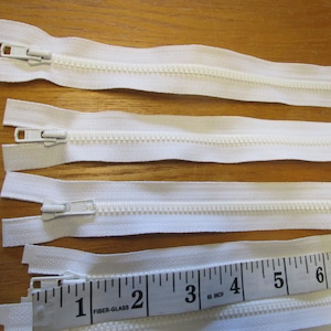 Puede incluir: Cuatro cierres de cremallera blancos con un deslizador de plástico blanco. Las cremalleras están dispuestas en una fila sobre una superficie de madera. Una regla con marcas negras es parcialmente visible en la esquina inferior derecha de la imagen. La regla está marcada en pulgadas.