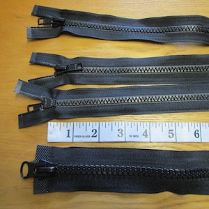 May include: Several black zippers of varying lengths are displayed on a wooden surface, alongside a measuring tape. The zippers have silver teeth and black pulls. The measuring tape shows inches.