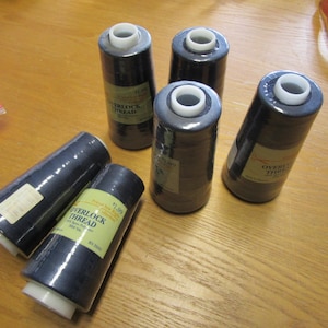 May include: Several spools of navy blue overlock thread are arranged on a wooden surface. Each spool has a white top and a label with text. A measuring tape is visible in the background.