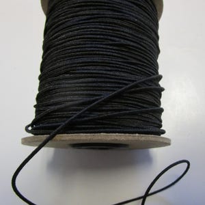 May include: A spool of black elastic cord, wound around a cardboard spool. The cord is thin and flexible, and is ideal for crafting and sewing projects.