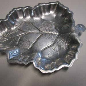 May include: A silver-colored leaf-shaped serving dish with embossed floral and leaf details. The dish has a scalloped edge and a small handle. This decorative tray is suitable for serving snacks or as a decorative accent.