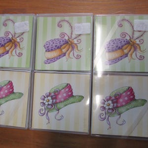 May include: Six square cards in clear plastic cases. The top row features a purple and orange hat with a bow and curly accents on a green striped background. The bottom row shows a green hat with a pink polka-dot ribbon and a white flower.