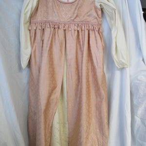 May include: A light pink costume with a diamond pattern, long sleeves, and a cream-colored bodice. The costume has a pink zipper, pink rope trim, and small tassels. The sleeves are cream-colored and long.