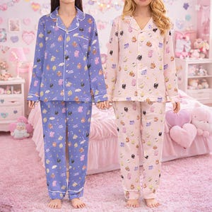 May include: Two sets of pajamas, one lavender and one pink, each with a button-down top and pants. The pajamas are patterned with cartoon characters, clouds, and moons. White trim accents the collar, cuffs, and pockets.
