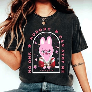 May include: Dark gray t-shirt with a pink bunny graphic inside a white arch, surrounded by the text "NO ONE CAN STOP ME". The bunny wears a jacket and boxing gloves. The name "changbin" is below the bunny.