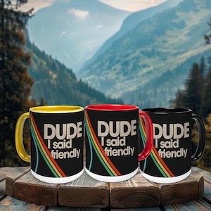 Könnte beinhalten: Drei schwarze Keramikbecher mit dem weißen Text "DUDE I said friendly" und einem Retro-Regenbogen-Design. Ein Becher hat einen gelben Henkel und Innenraum, einer einen roten, und einer einen schwarzen.