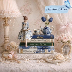 May include: A decorative figurine of a woman reading, perched on a stack of books. The books are titled "Victoria", "Jane Eyre", "Wuthering Heights", and "Little Women". A blue and white teapot and teacup are nearby, with a vase of tulips. A hand mirror and pearls complete the scene.