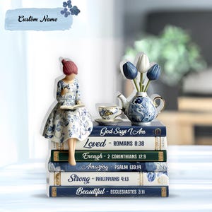 May include: A decorative figurine of a woman reading, perched atop a stack of books. The books are inscribed with inspirational words. A teapot with blue and white floral design and a teacup with the name "Lisa" are also present. The text "Custom Name" is on a light blue banner.