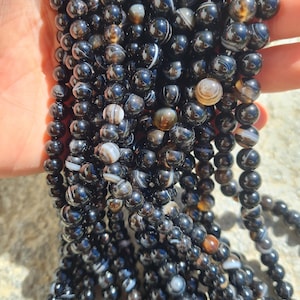 Può includere: Fili di perline di agata nera con fasce bianche e marroni. Le perline sono rotonde e lucide, riflettendo la luce. Le perline sono infilate, pronte per la creazione di gioielli o altri lavori artigianali.