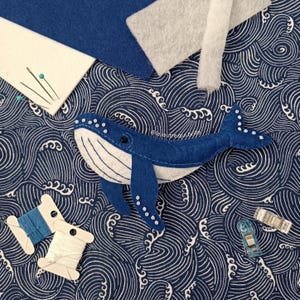 Könnte beinhalten: Ein handgefertigter Wal aus blauem Filz mit weißen Akzenten, platziert auf einem Stoff mit marineblauem und weißem Wellenmuster. Nähzubehör, darunter Garnrollen, Nadeln und Stoffreste, sind um den Wal herum angeordnet.