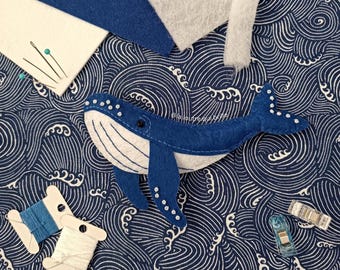 Può includere: Una balena di feltro blu fatta a mano con dettagli bianchi, posizionata su un tessuto a fantasia a onde blu navy e bianco. Forniture per cucito, tra cui rocchetti di filo, spilli e ritagli di tessuto, sono disposte attorno alla balena.