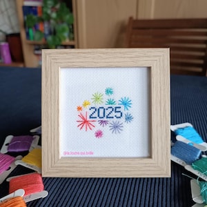 May include: A framed cross-stitch piece with the year 2025 in blue, surrounded by colorful starbursts. The frame is light wood. The text "@la.loutre.qui.brille" is at the bottom.